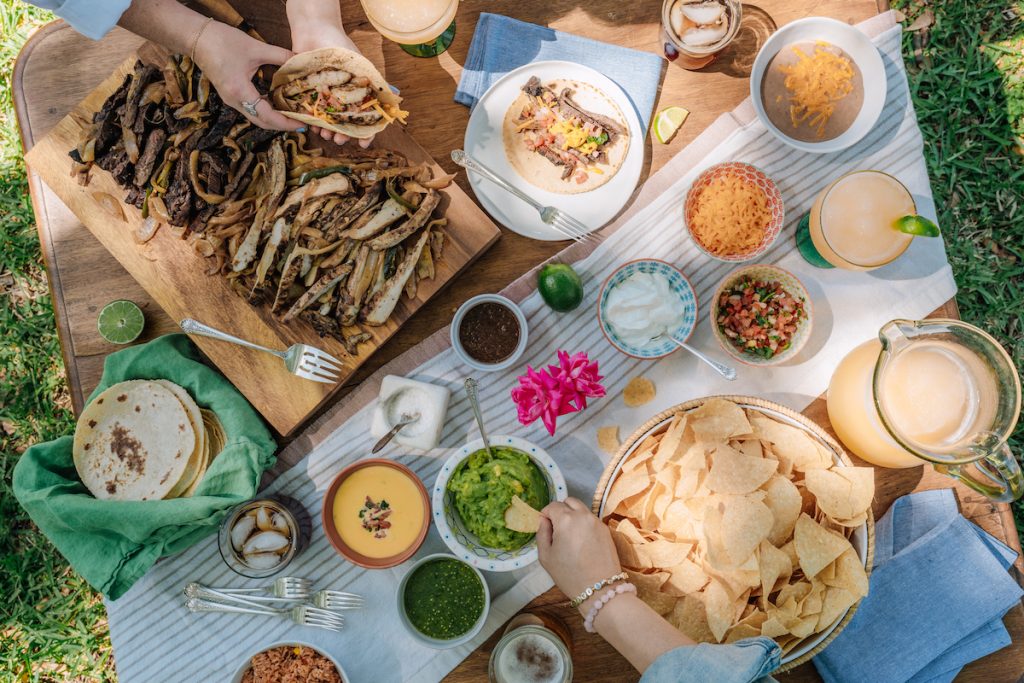 Food display with guacamole, chips, someone making a fajita, spicy sliced beef, queso, and a margarita pitcher.