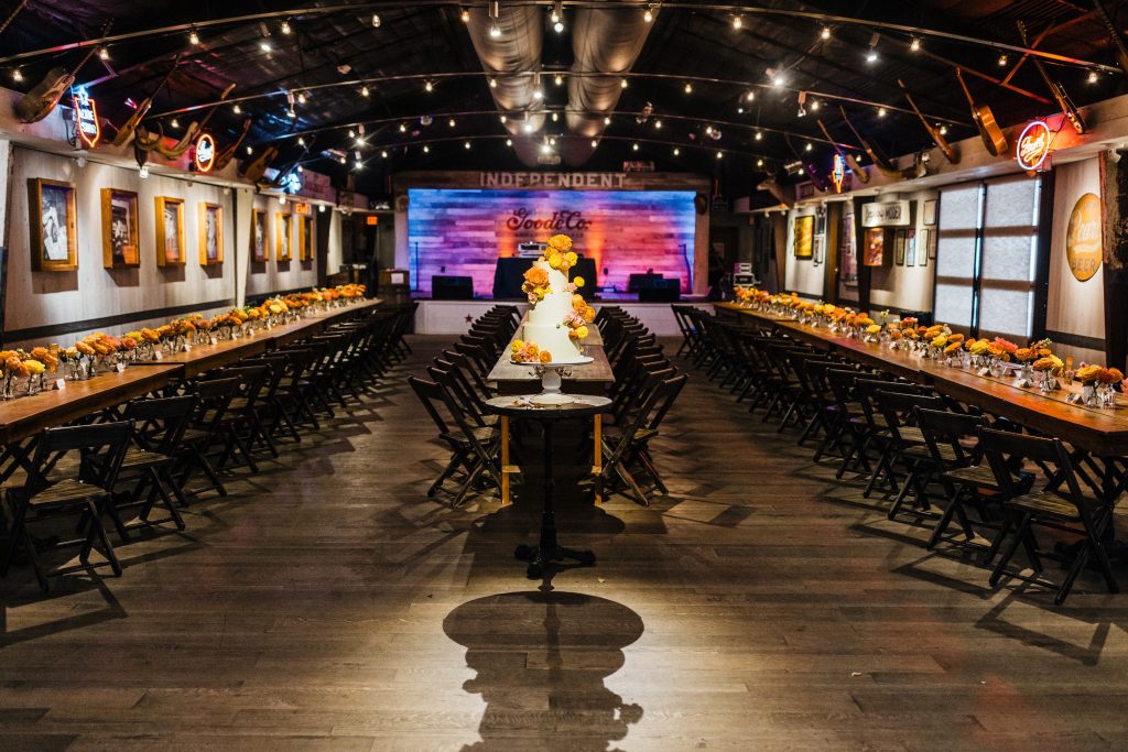 Wide shot of wedding venue with white tiered cake garnished with orange flowers in the middle, and long brown tables on either side