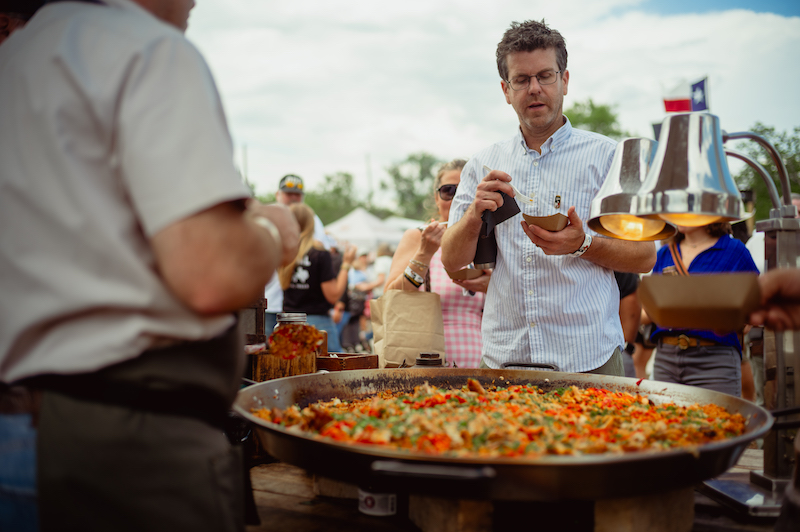 Big frying pan full of paella with a customer with eyeglasses sampling a small portion.