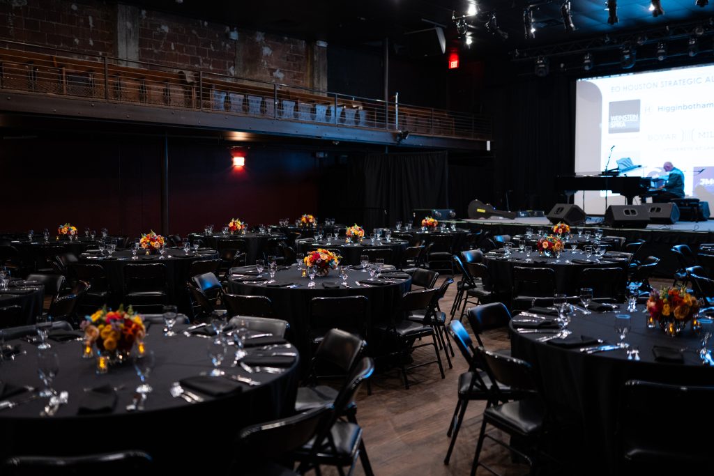 Large catering room with round tables, black cloths, flower centerpieces, and cutlery. 