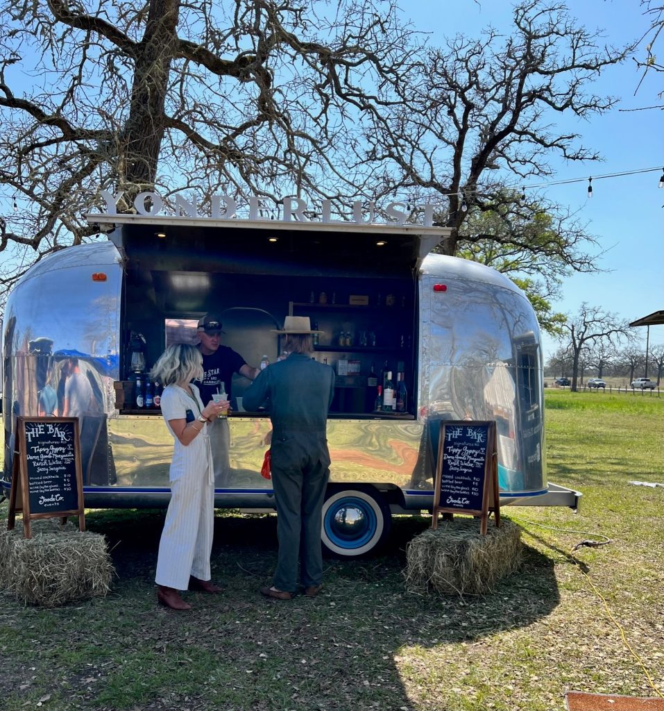 The yonderlust camper with its side window open, serving drinks to a couple under a large tree at the Round Top event.