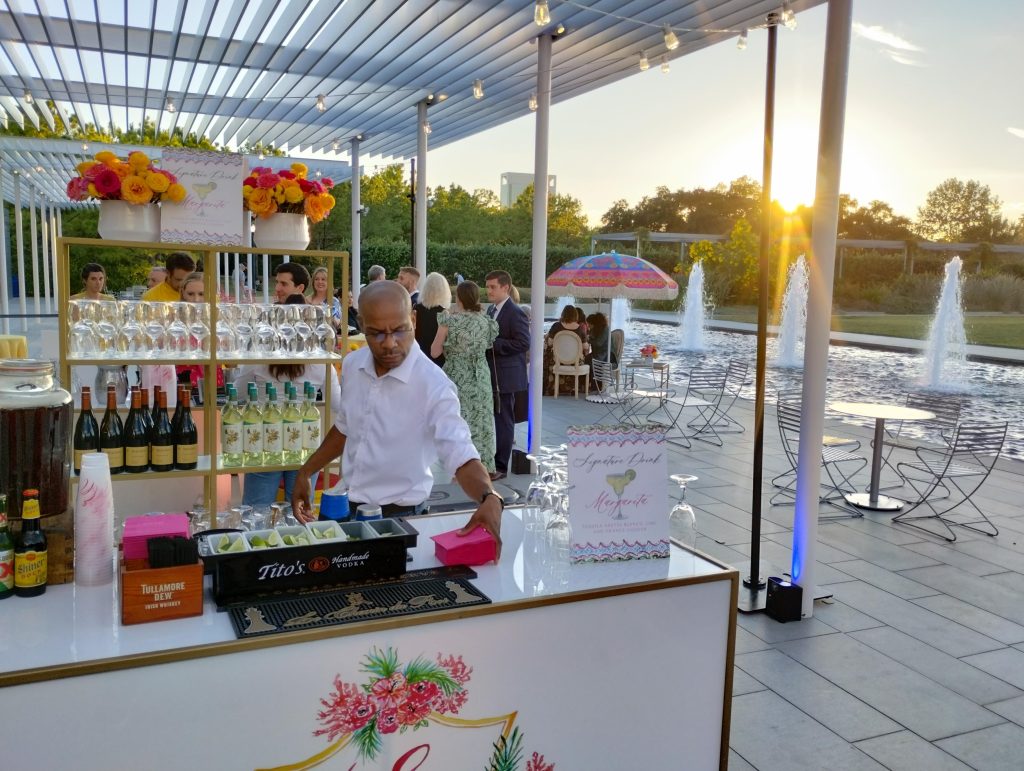 A white-shirted bartender arranging napkins at his outdoor bar, with the sun setting in the background, next to a fountain pool.