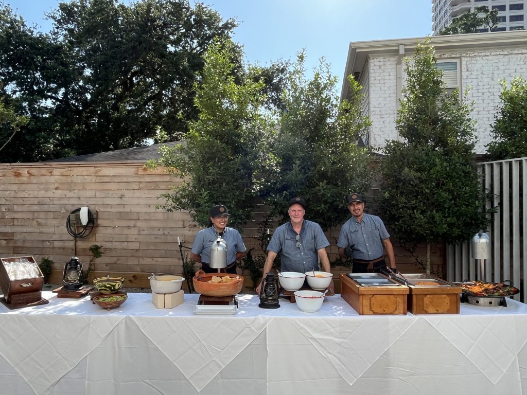 Three grey-shirted workers standing behind a catering table in a private backyard with a table full of food, ready to serve.